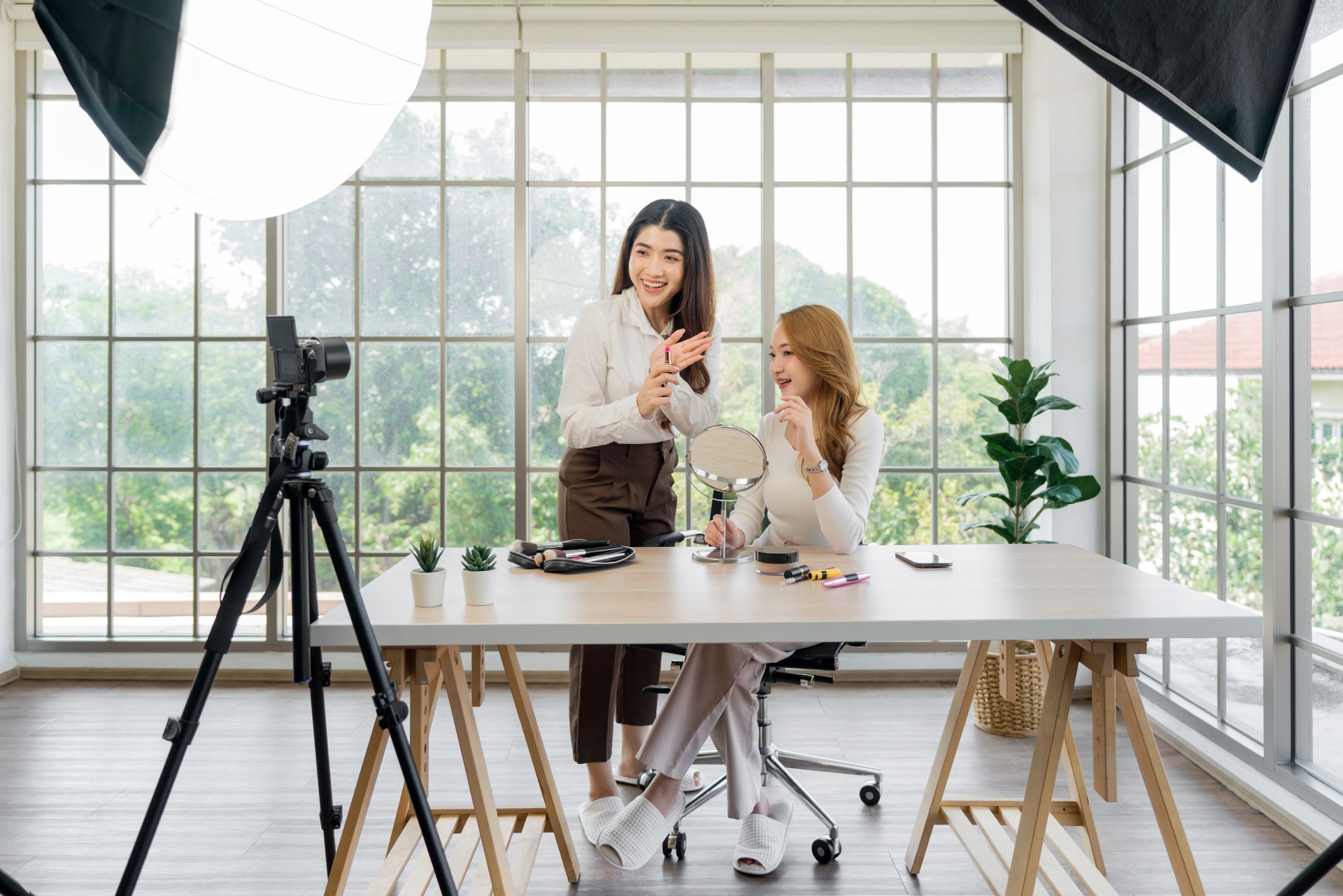 Two asian women creating content for social media. They are using a studio setup that include a camera mounted on a tripod, lighting equipment and various makeup product on the table.